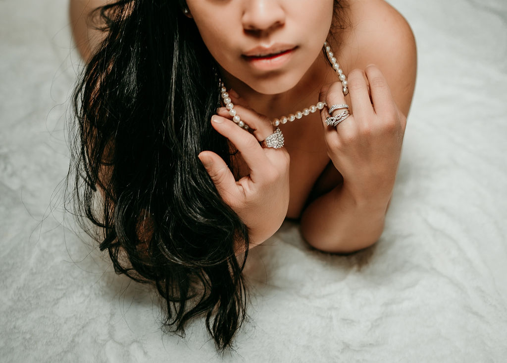 A woman lays on a white blanket holding her pearl necklace with multiple diamond rings annapolis nail salons