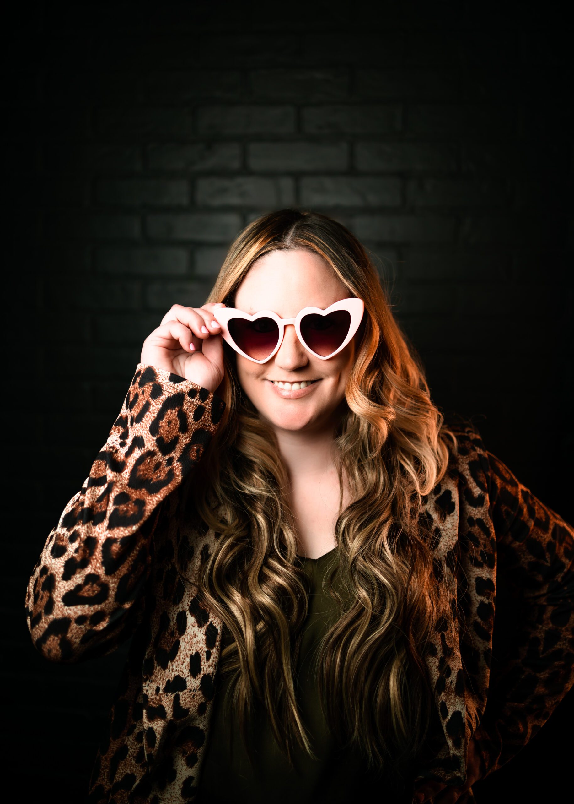 professional headshot of woman with blonde hair smiling and wearing pink heart sunglasses against black backdrop